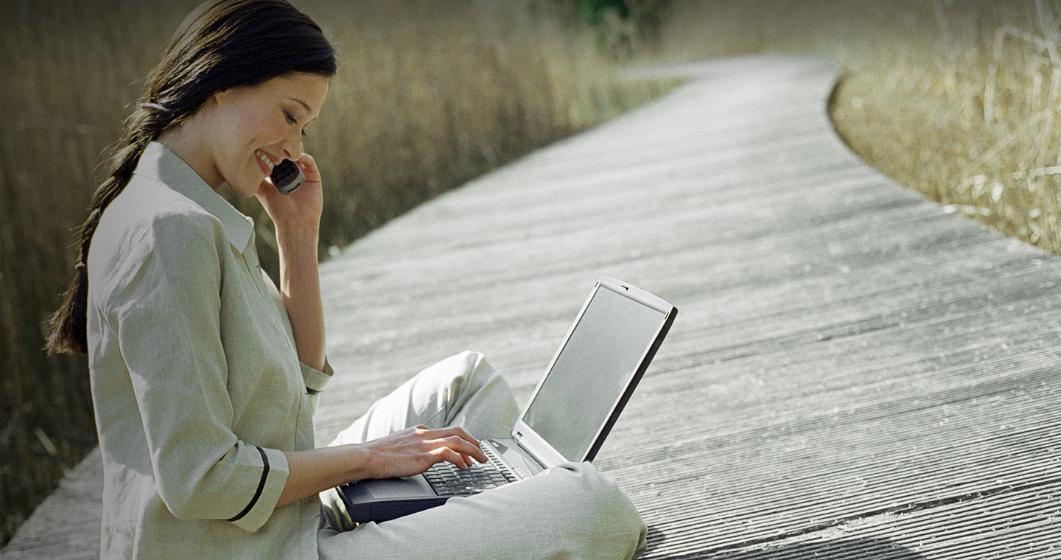 Lady smiling, while sitting cross-legged, using a mobile phone and laptop