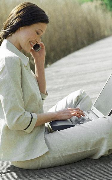 Lady smiling, while sitting cross-legged, using a mobile phone and laptop